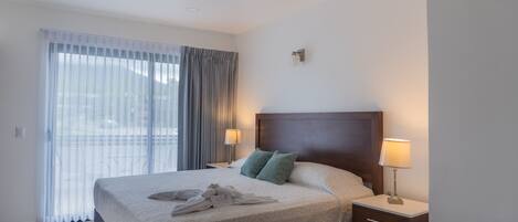 A hotel room with a large bed, two bedside lamps, a nightstand, and a view through sheer curtains.