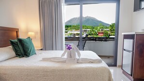 Quarto de hotel com cama, almofadas, toalha, mesa de cabeceira, luminária e varanda com vista para a montanha e paisagem urbana.