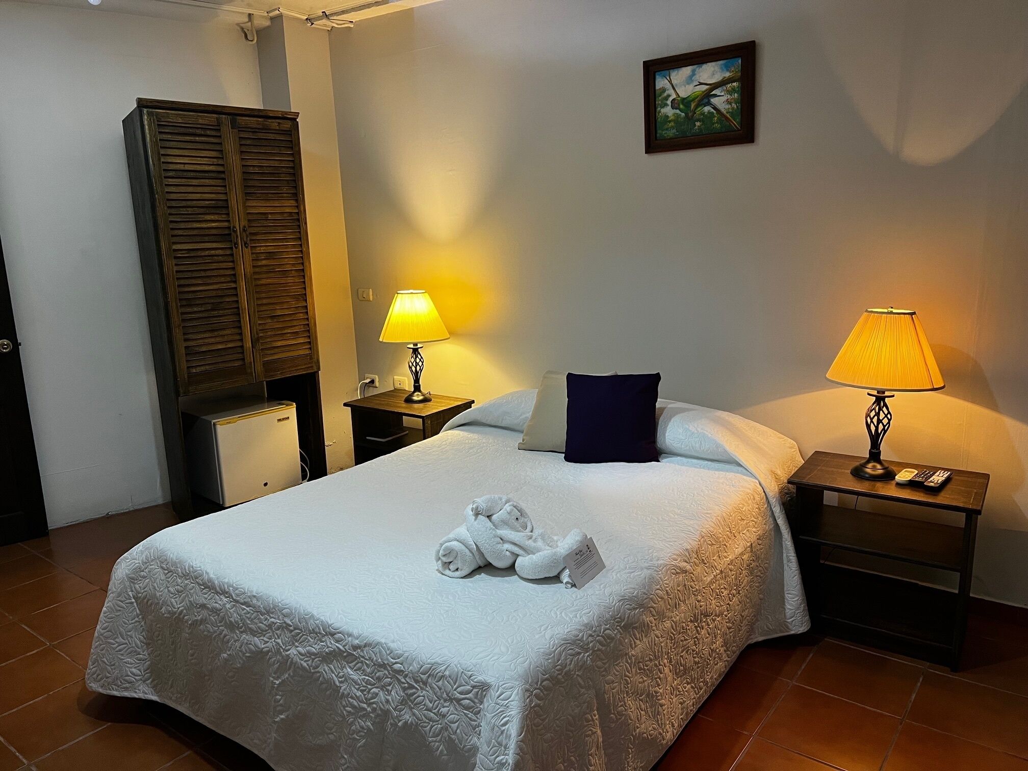 A neatly made bed with a white duvet, two bedside tables with lamps, a wooden wardrobe, and a painting on the wall.