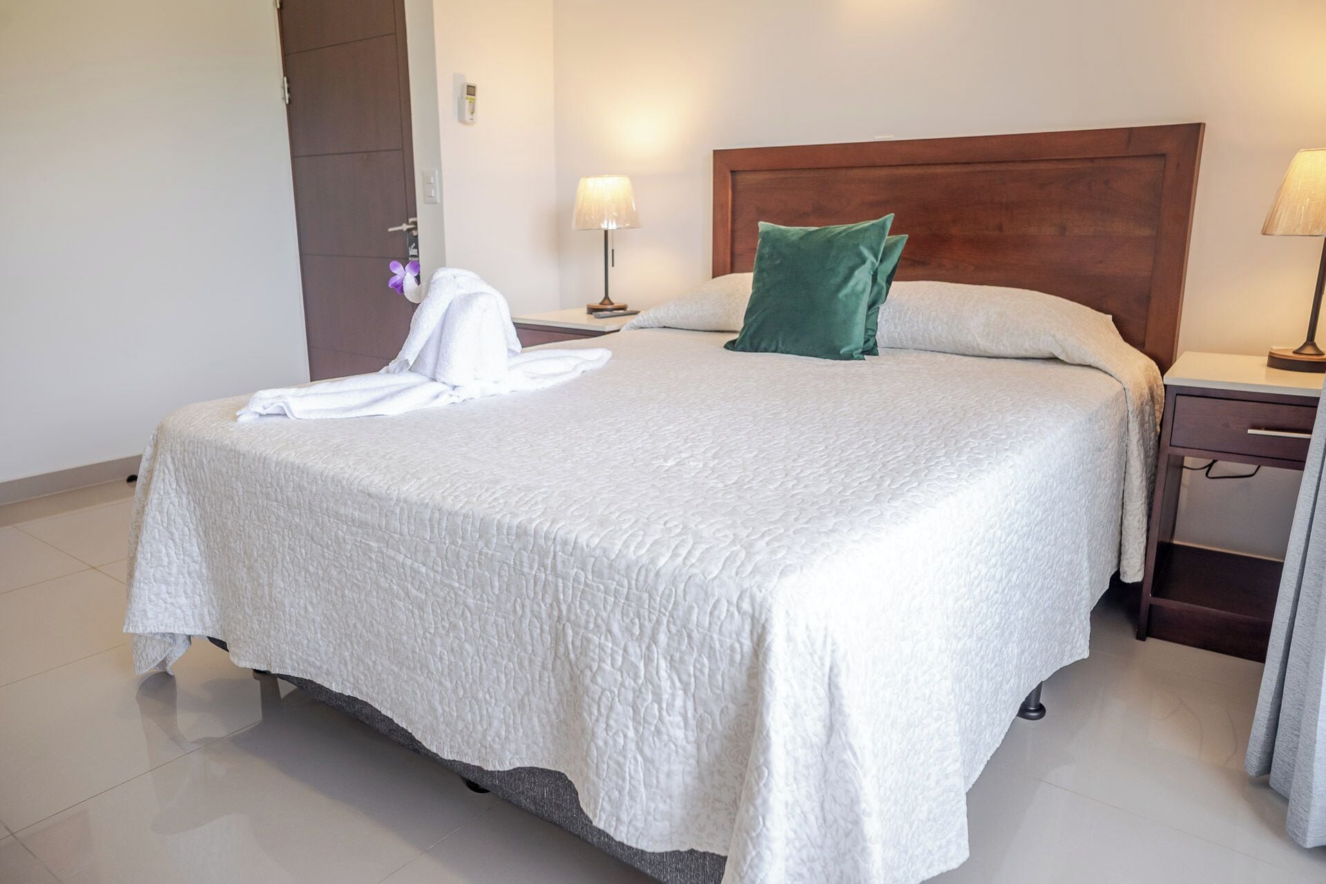 Una cama bien hecha con edredón blanco, almohada verde y bata blanca al lado. Hay dos mesitas de noche con lámparas, cabecero de madera y una puerta a la izquierda.