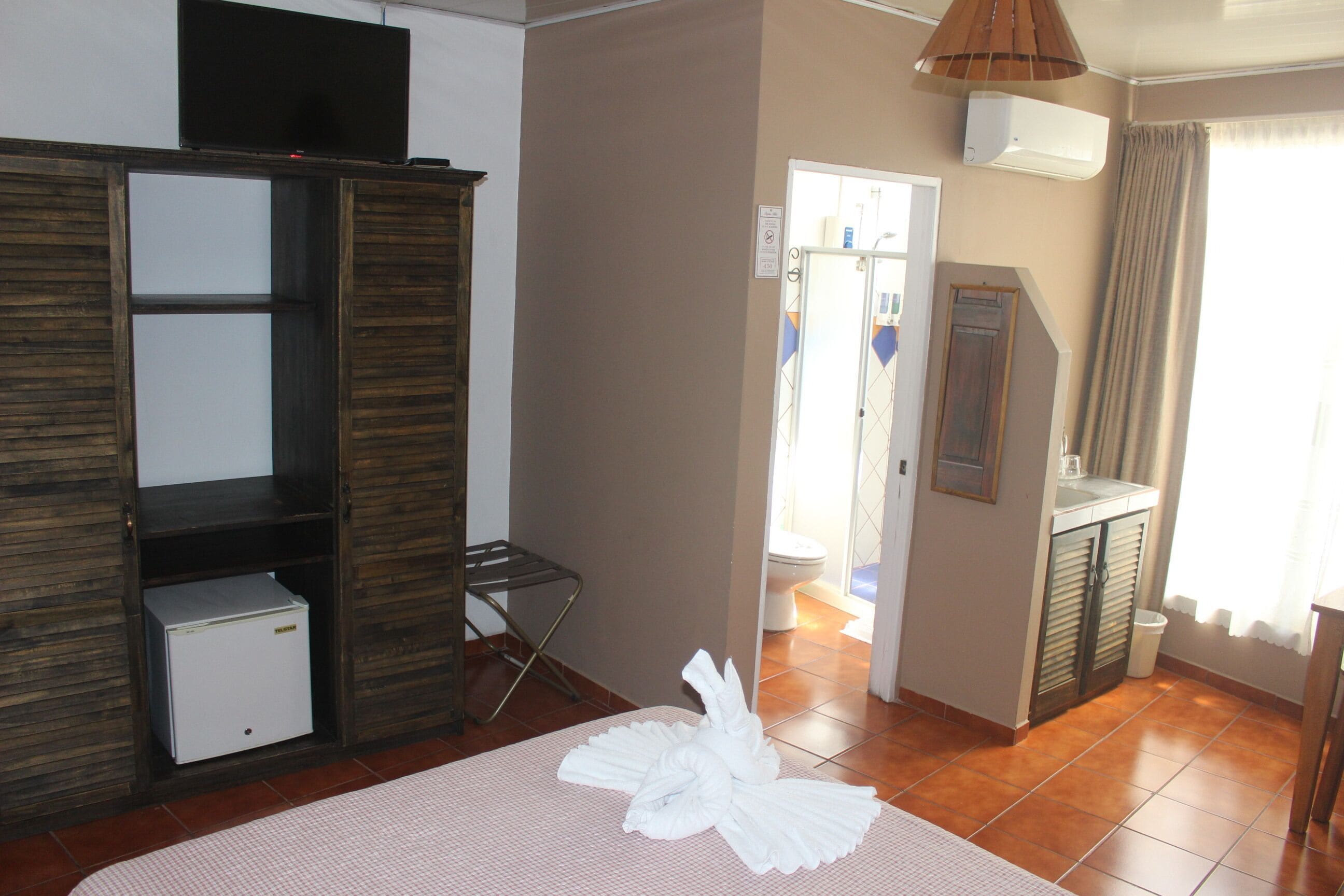 A hotel room with a bed, a wardrobe, a small fridge, a bathroom visible through an open door, and a hanging light fixture.