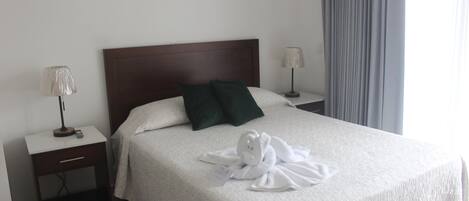 Un lit bien fait, recouvert d’une courtepointe blanche, avec deux oreillers vert foncé et une serviette blanche posée sur le lit. Il y a deux tables de chevet, chacune avec une lampe.