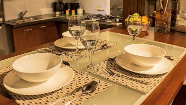 Una mesa de comedor dispuesta con platos, tazones y cubiertos blancos, y una servilleta debajo.
