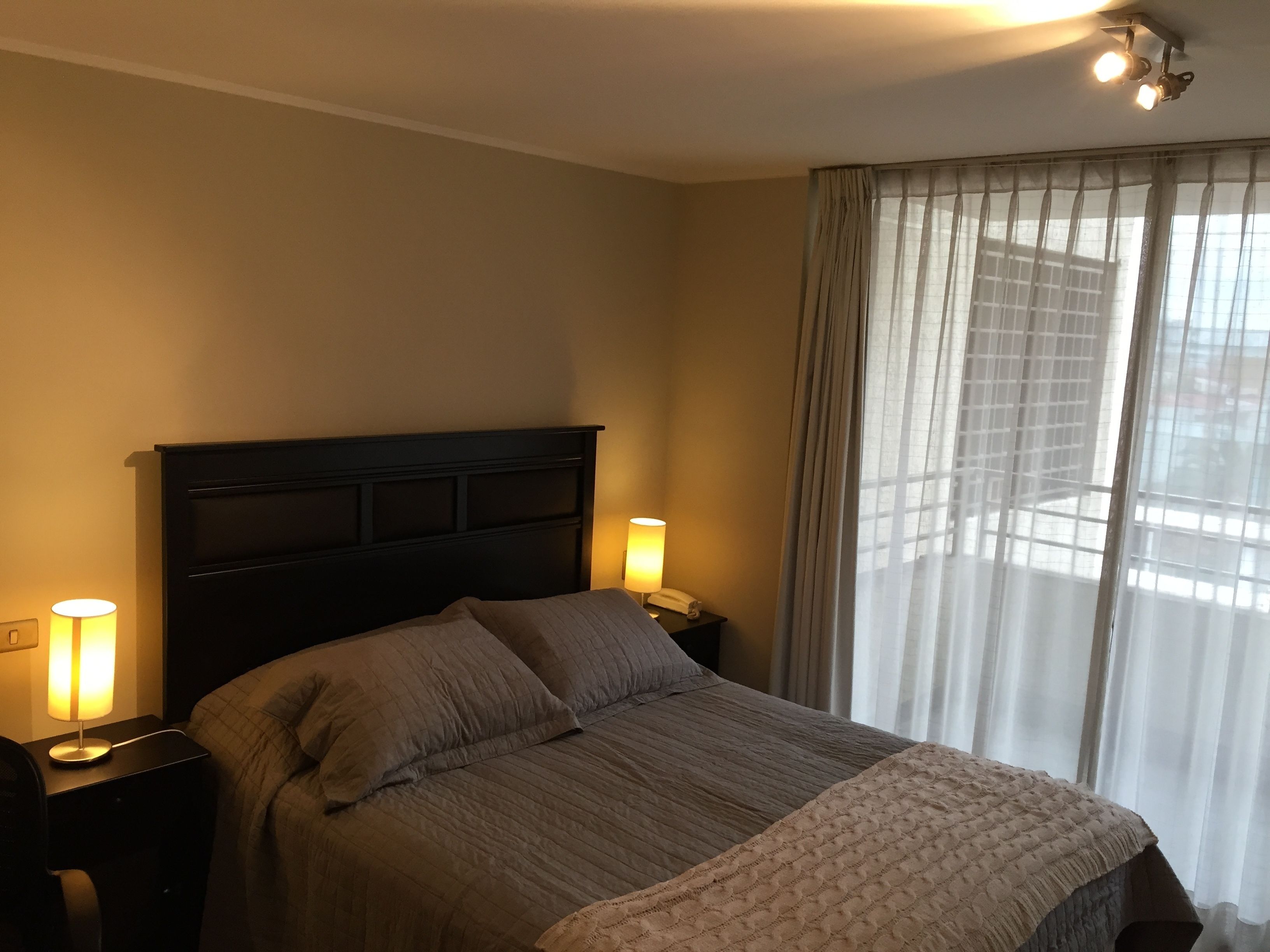 Ein Hotelzimmer mit einem großen Bett, zwei Nachttischlampen, einem Nachttisch und einem Fenster mit durchsichtigen Vorhängen.