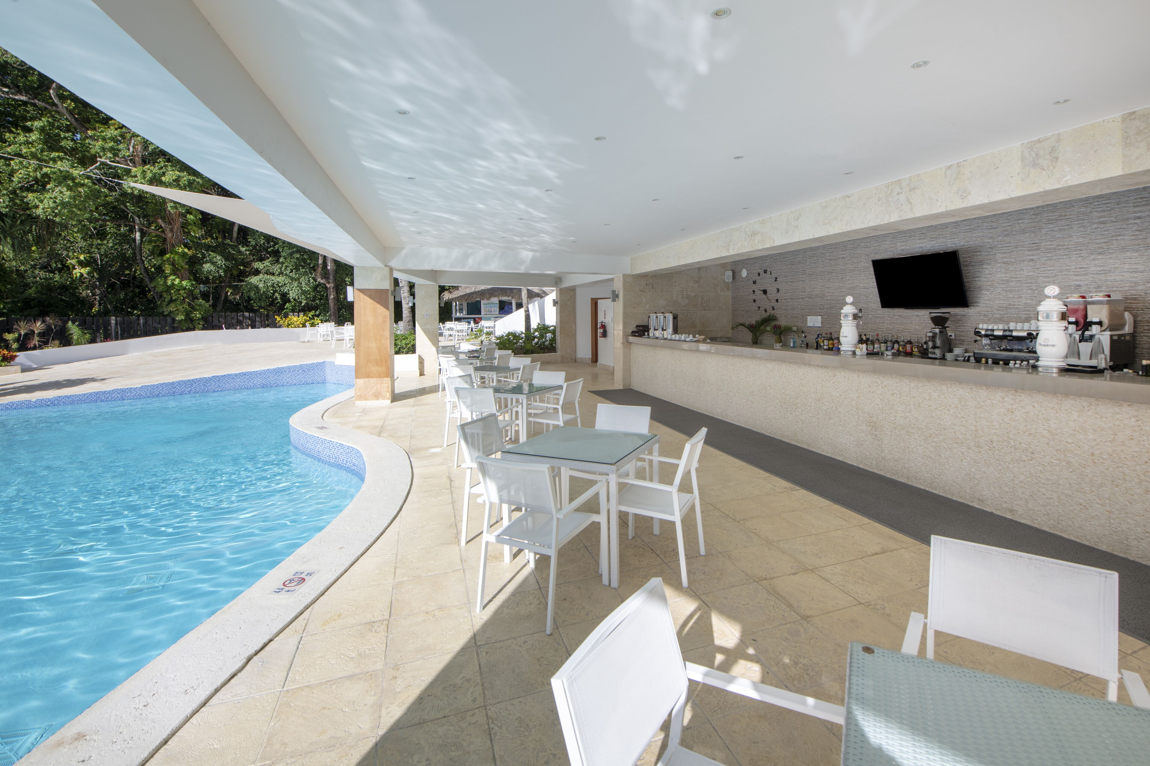Un área de piscina al aire libre con sillas y mesas blancas, un bar con televisor de pantalla plana y suelo de baldosas.