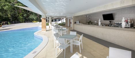 Un área de piscina al aire libre con sillas y mesas blancas, un bar con televisor de pantalla plana y suelo de baldosas.