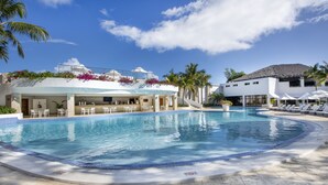 A swimming pool with lounge chairs and tables, surrounded by palm trees and a building with a white facade.