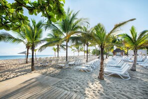 Une plage bordée de rangées de chaises longues blanches et de palmiers, avec une terrasse en bois et une vue sur l’océan.