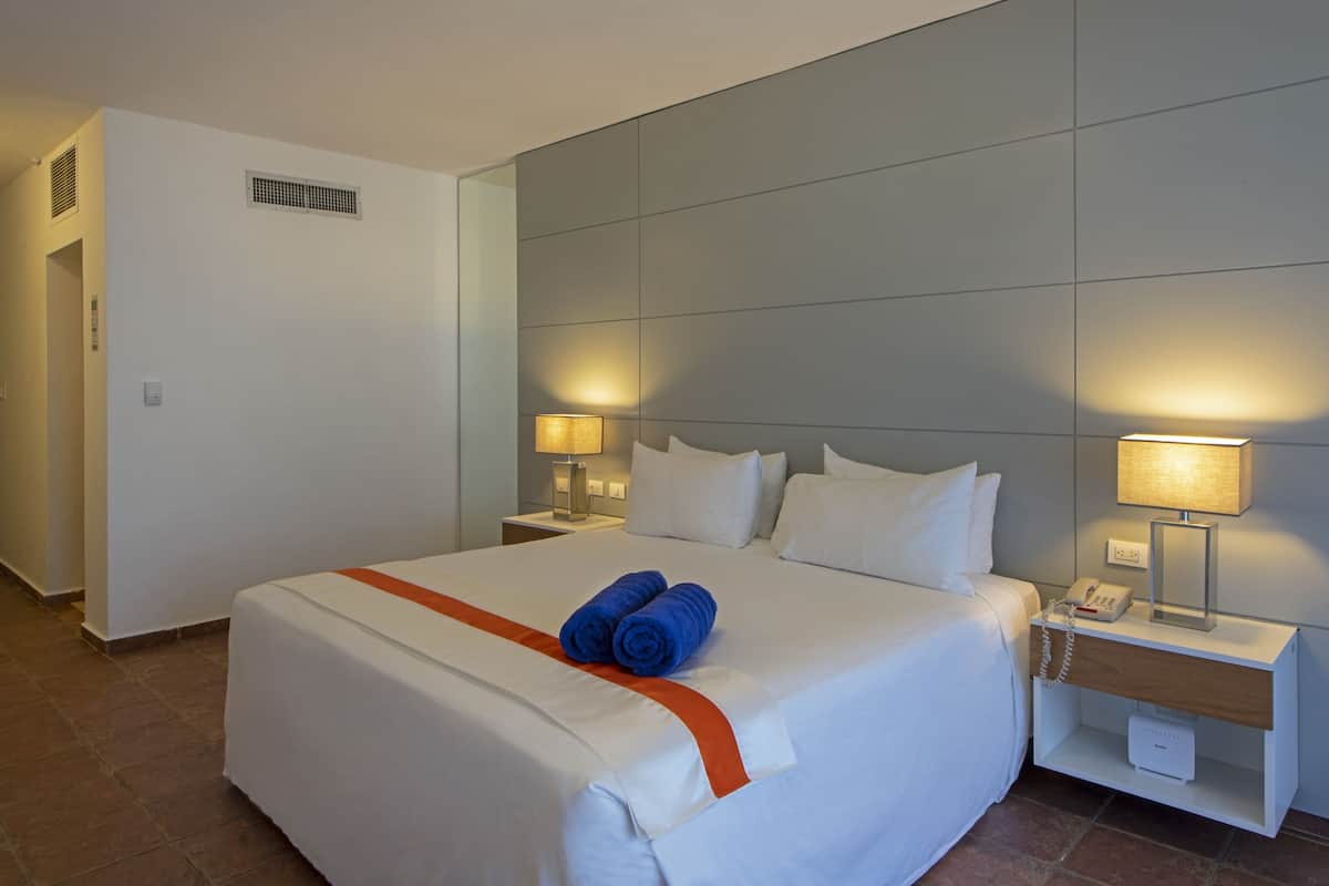 Une chambre d’hôtel avec un grand lit, deux lampes de chevet, une table de chevet avec un téléphone et une lampe, et une bouche d’aération fixée au mur.