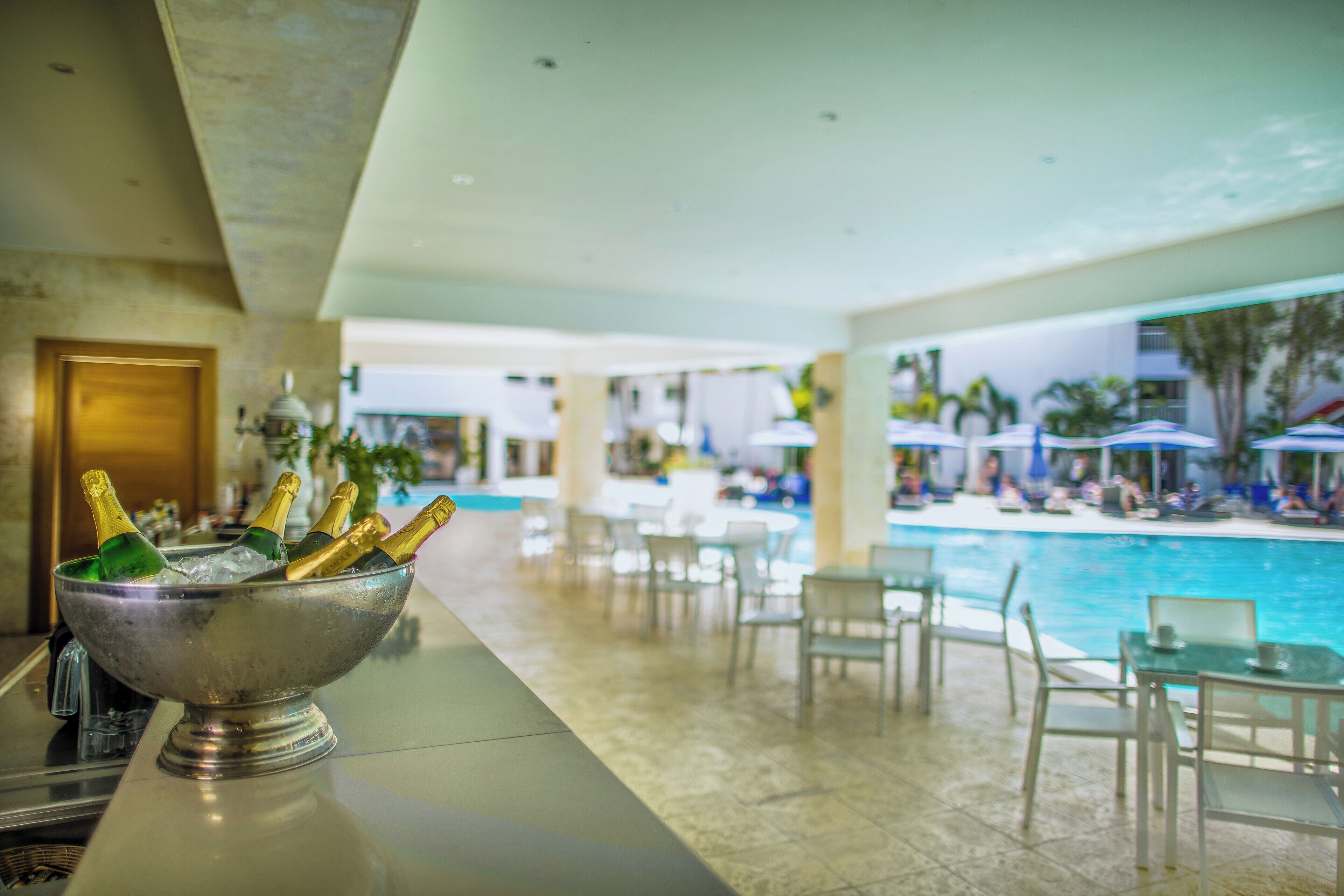 Un espace bar avec un récipient en métal contenant des bouteilles de champagne, un coin salon au bord de la piscine avec des tables et des chaises, et une vue sur la piscine.