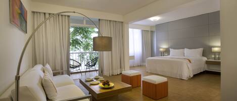 A modern hotel room with a large bed, a sofa, a coffee table, and a dining table. There is a window with curtains, a lamp, and a painting on the wall.
