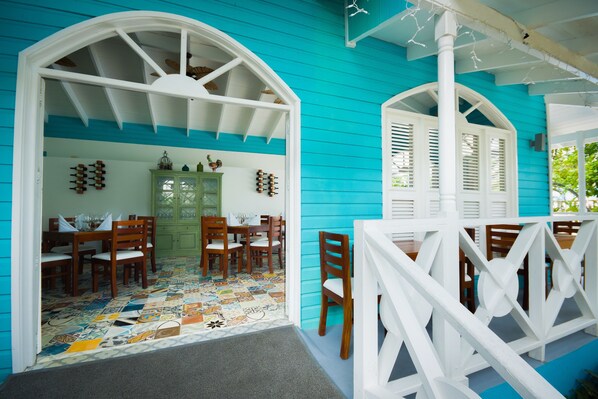 Exterior azul brillante con detalles arquitectónicos blancos, un comedor con muebles de madera y un piso de baldosas coloridas.
