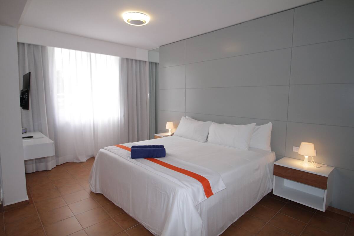 Une chambre d’hôtel avec un grand lit, une table de chevet avec une lampe, un téléviseur fixé au mur et une fenêtre avec des rideaux.