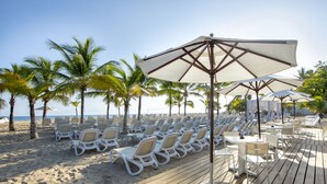 A beachside resort with rows of white lounge chairs and umbrellas, palm trees, and a wooden deck.