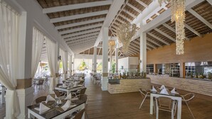 A spacious restaurant with high ceilings, wooden beams, and a central bar area. Tables are set with white tablecloths and chairs.