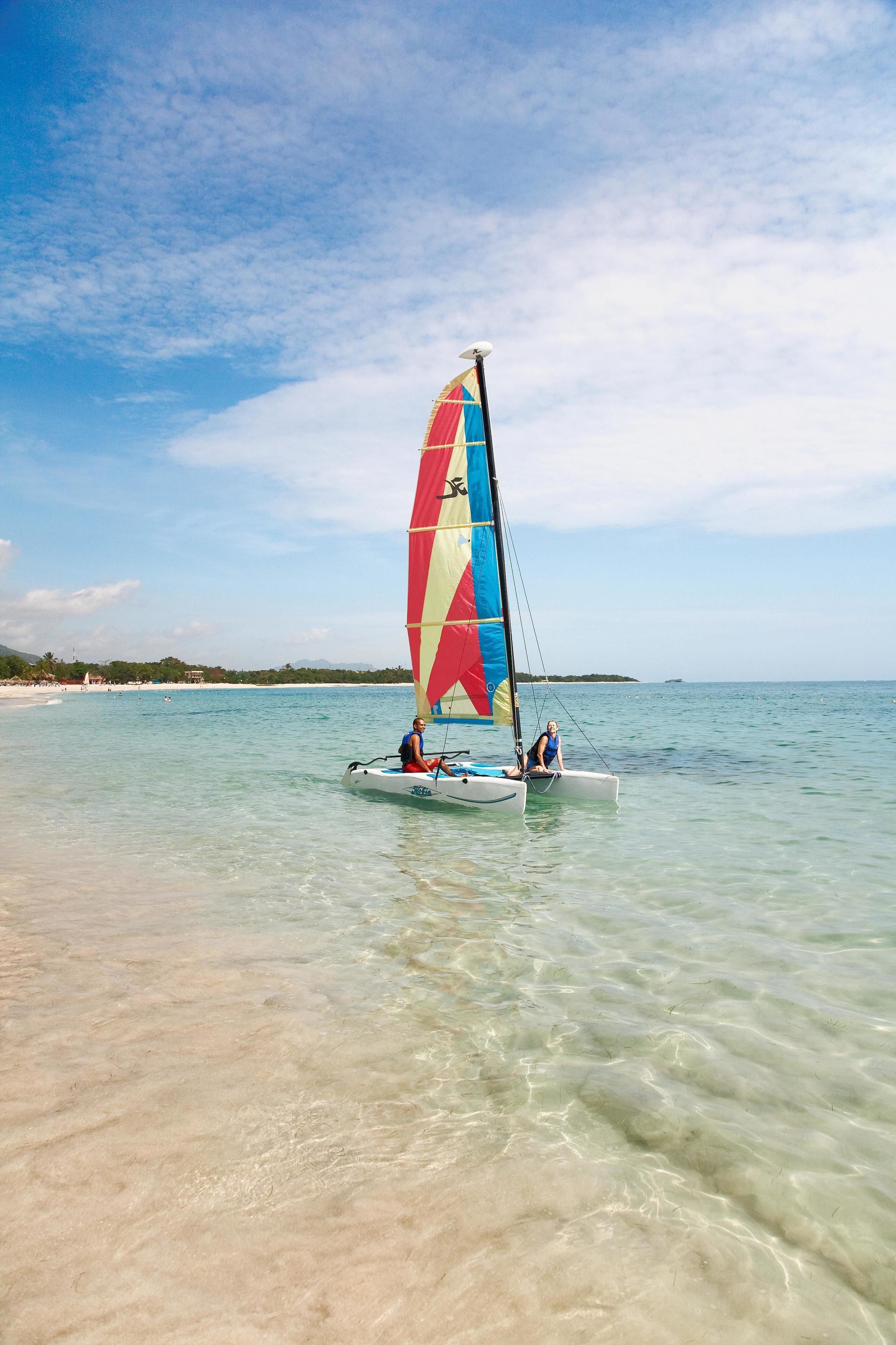 A sailboat with a colorful sail on clear turquoise water near a sandy beach.