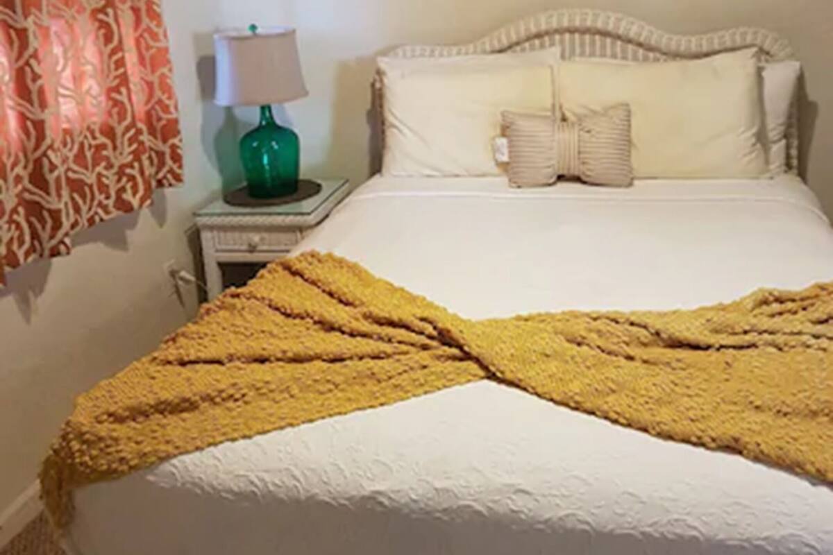 Una cama bien tendida con un edredón amarillo, una lámpara de noche y un cuadro en la pared.