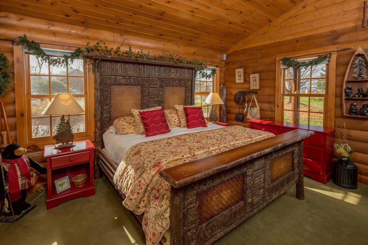 A cozy log cabin bedroom with a large wooden bed, two bedside tables, a dresser, and a window with a view of the outdoors.