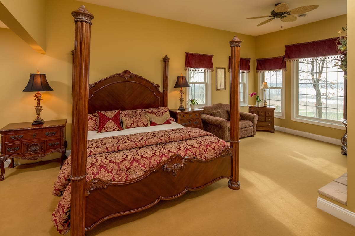 A four-poster bed with a wooden frame and red bedding, a wooden nightstand with a lamp, a chair, and a window with a view of trees.