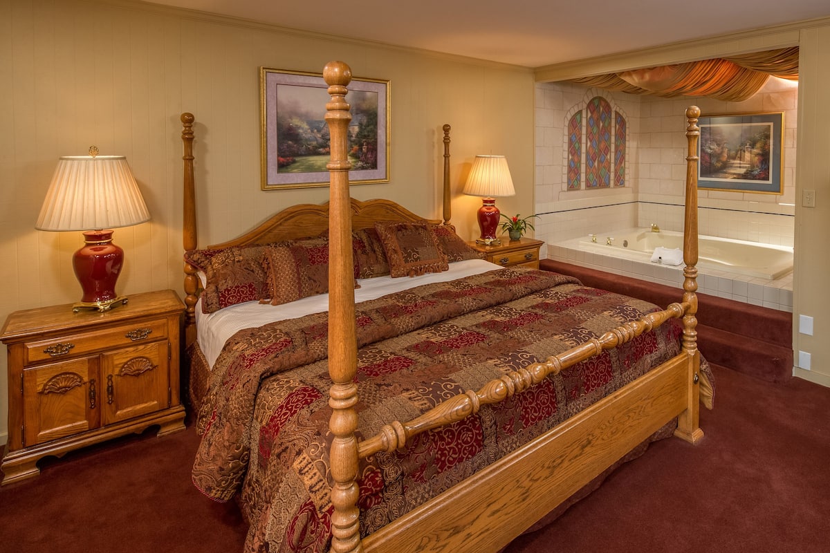 A four-poster bed with a red and brown patterned bedspread, a wooden nightstand with a lamp, a wooden dresser, and a large window with stained glass.
