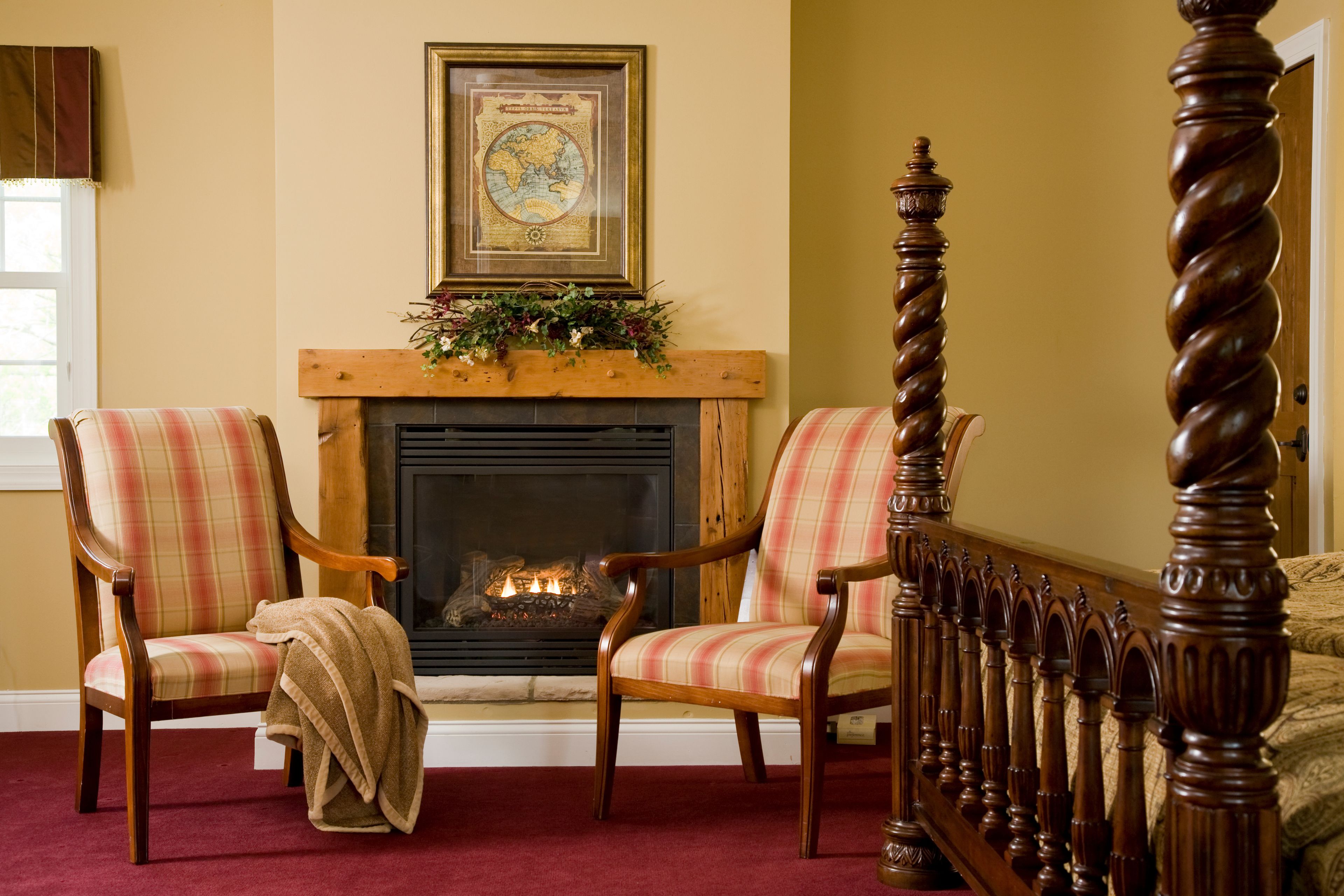 A room with a fireplace, two plaid chairs, and a four-poster bed.