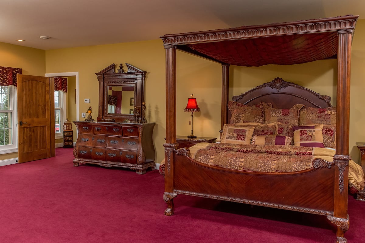 A four-poster bed with ornate carvings, a wooden dresser with multiple drawers, a mirror with a decorative frame, and a red carpeted floor.