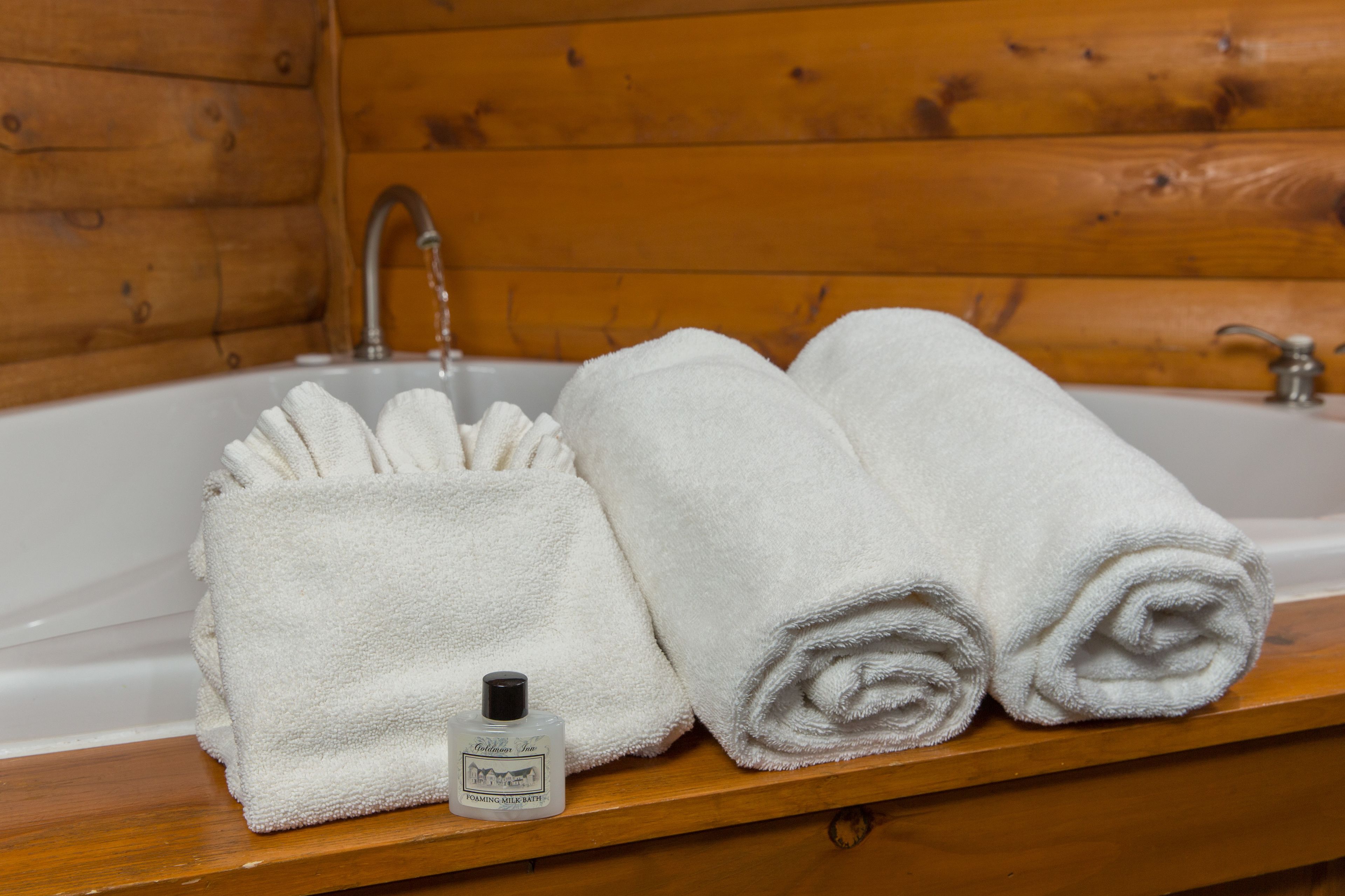 A wooden cabin bathroom with a white bathtub, two rolled white towels, and a bottle of bath soap on a wooden shelf.