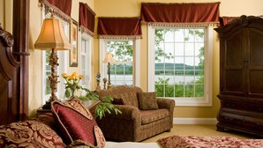 A room with a sofa, armchair, and a large wooden cabinet. There are two windows with curtains, and a lamp on a side table.