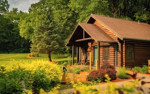 A log cabin with a porch, surrounded by a well-maintained garden and a forested area.