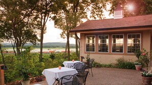 An outdoor patio with tables and chairs, surrounded by greenery and a view of the landscape.