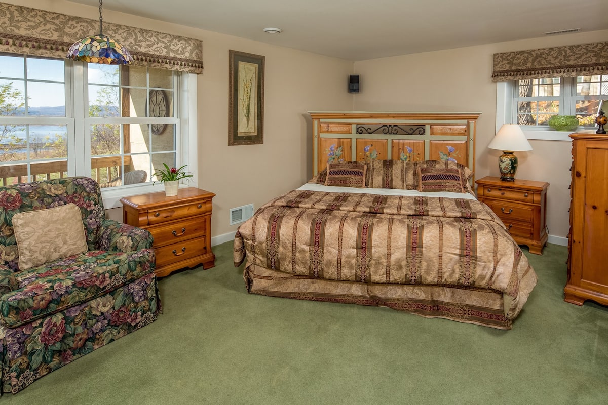 A bedroom with a floral-patterned sofa, a bed with a striped bedspread, a wooden nightstand, and a view of the outdoors through large windows.