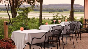 An outdoor dining area with tables and chairs, surrounded by trees and a view of a river.
