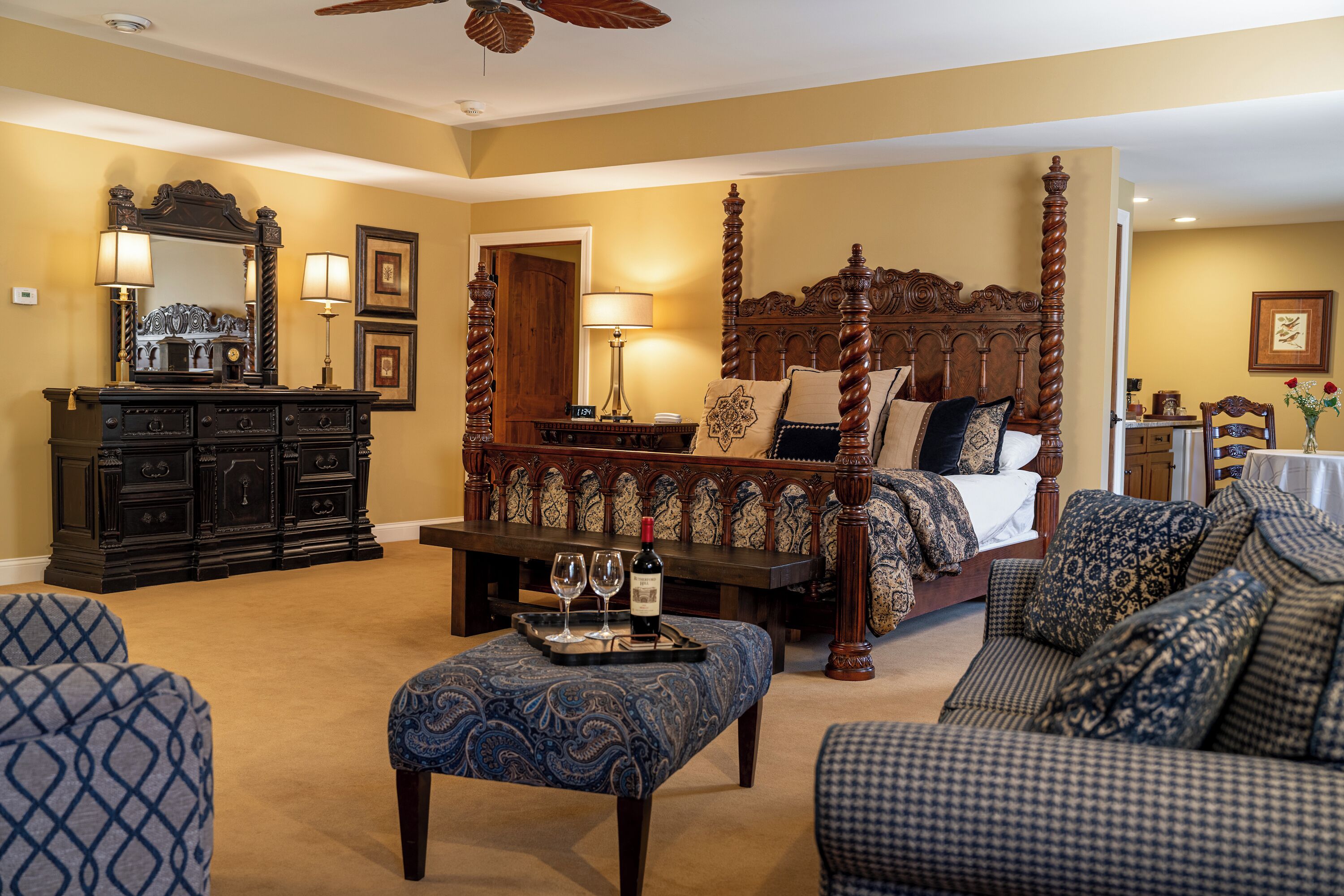 A spacious bedroom with a four-poster bed, a sitting area with patterned sofas, a wooden dresser, and a large mirror.