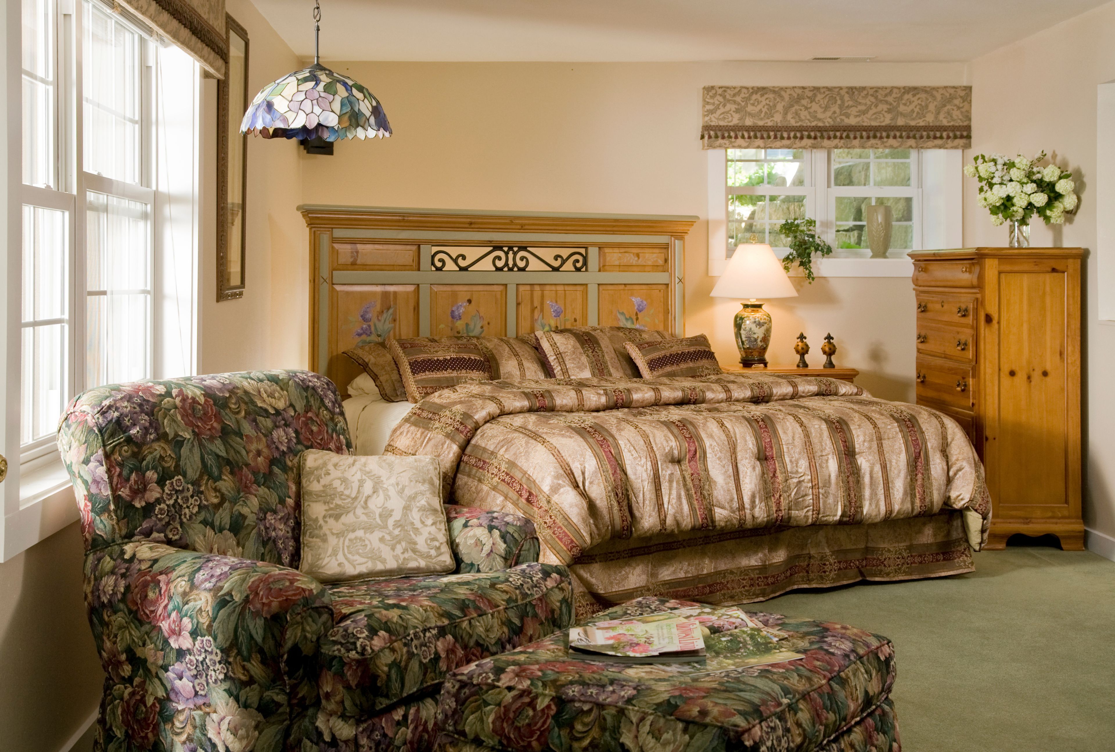 A bedroom with a floral-patterned armchair, a large bed with a striped bedspread, a wooden dresser, and a window with a view of greenery.