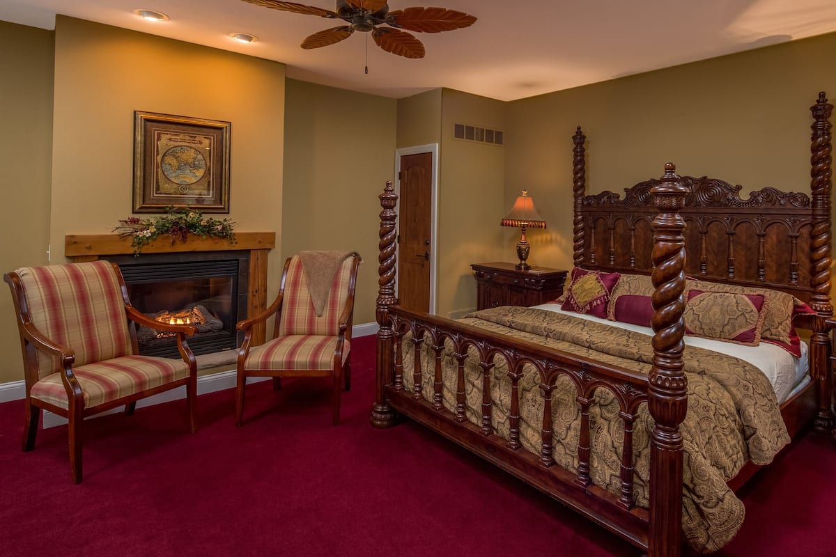 A bedroom with a four-poster bed, two armchairs, a fireplace, and two bedside lamps.