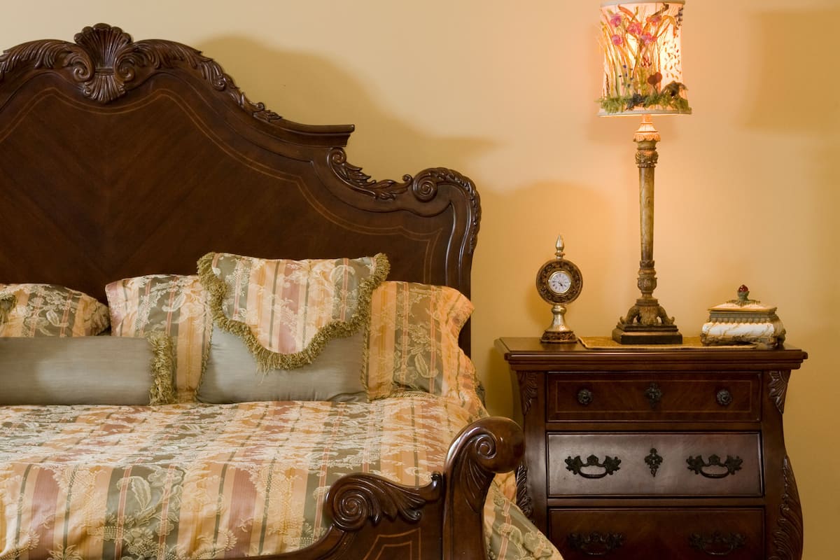 A traditional wooden bed with ornate carvings, a floral-patterned bedspread, and matching pillows. A wooden nightstand with a clock and a decorative lamp is beside the bed.