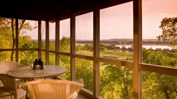 A covered outdoor seating area with wicker chairs and a round table, overlooking a scenic landscape with trees and a bridge. - Goldmoor Inn (Galena)