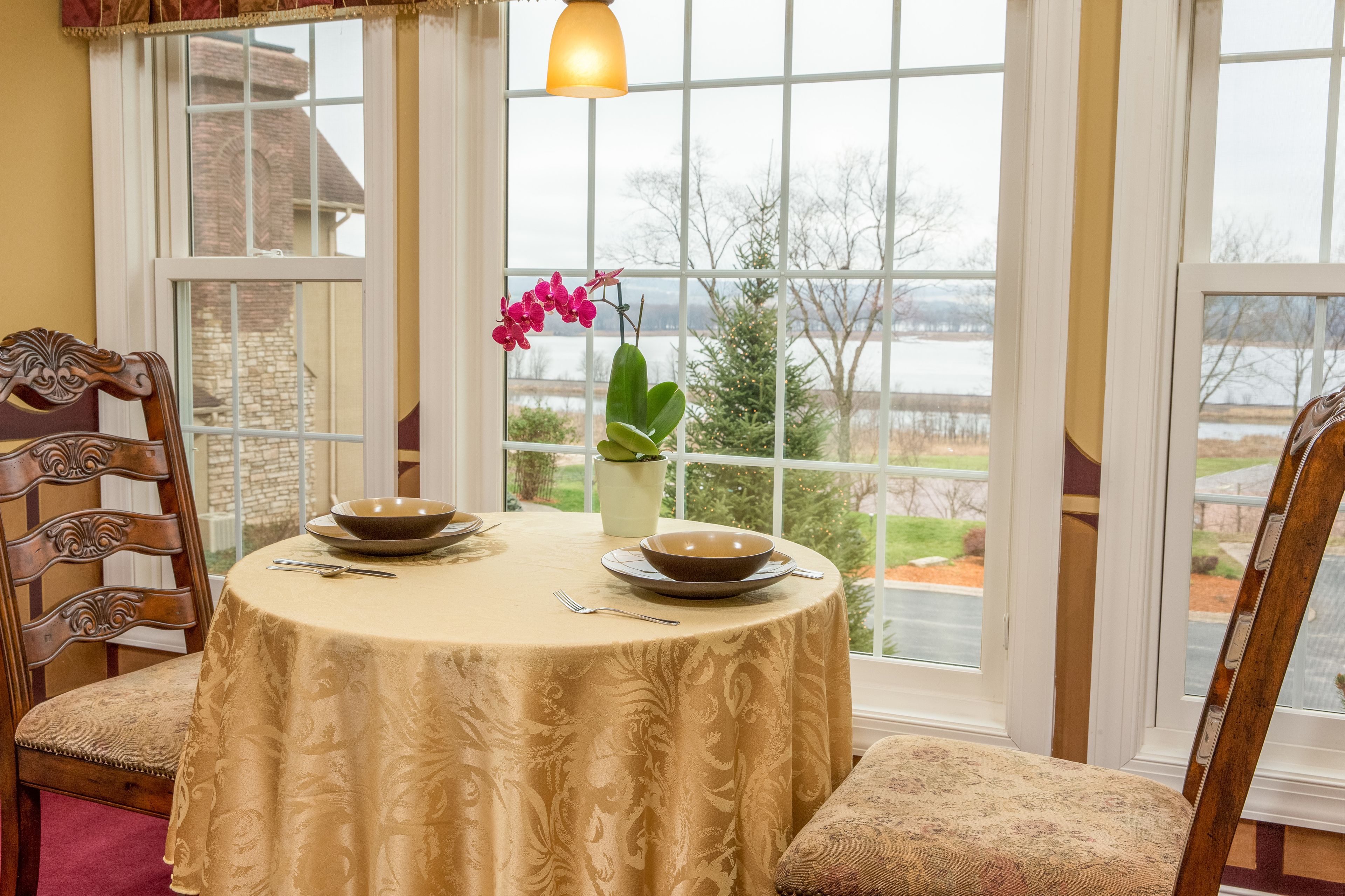 A dining area with a table set for two, a vase with flowers, and a view of a lake through large windows.