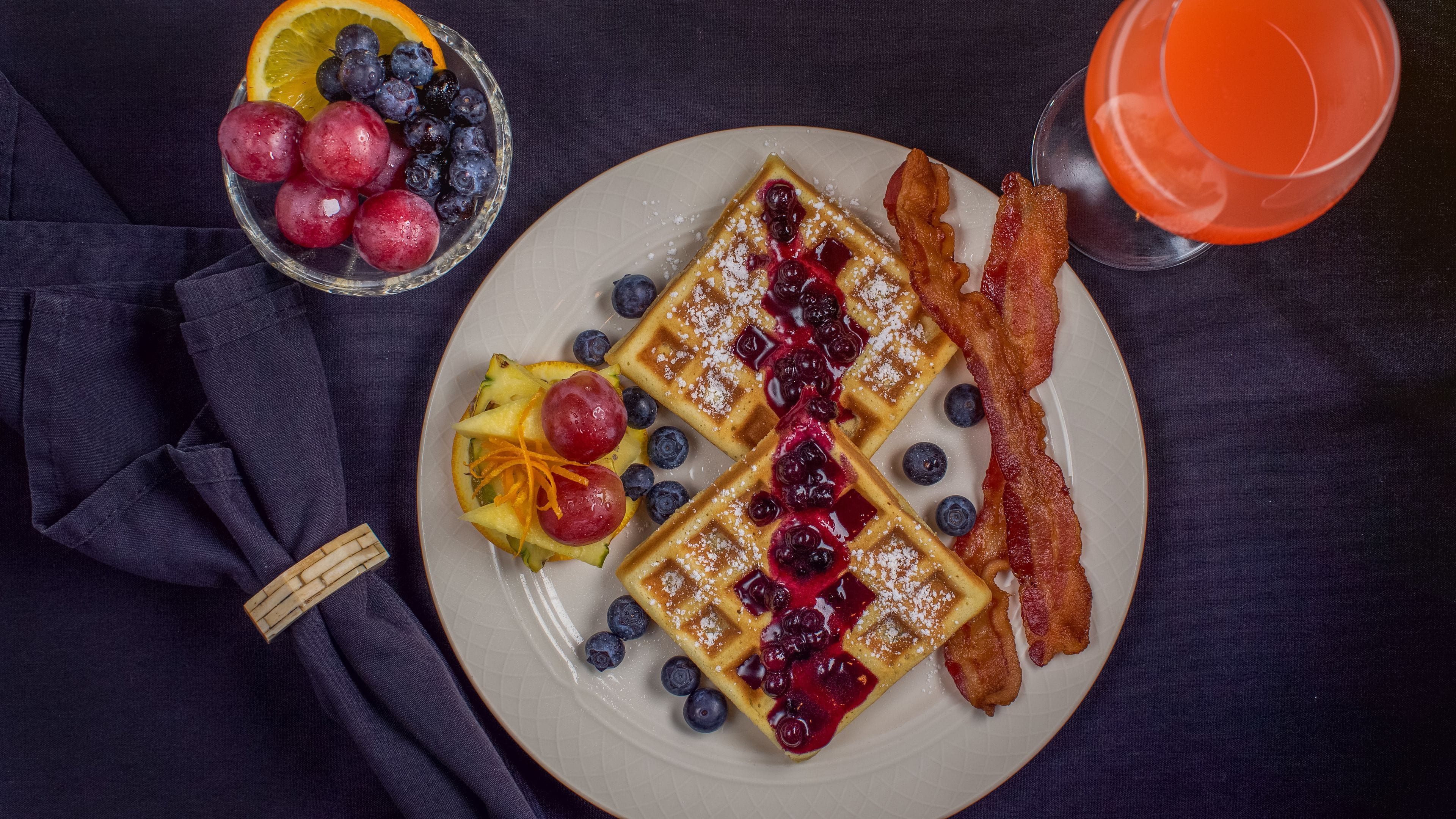 A plate with waffles, blueberries, and bacon, a bowl of fruit, and a glass of orange juice.