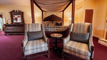 Una cama con dosel y cuatro columnas, con un edredón estampado, dos sillones tapizados con diseño geométrico, una mesita de madera y un aparador de madera con cajones.
