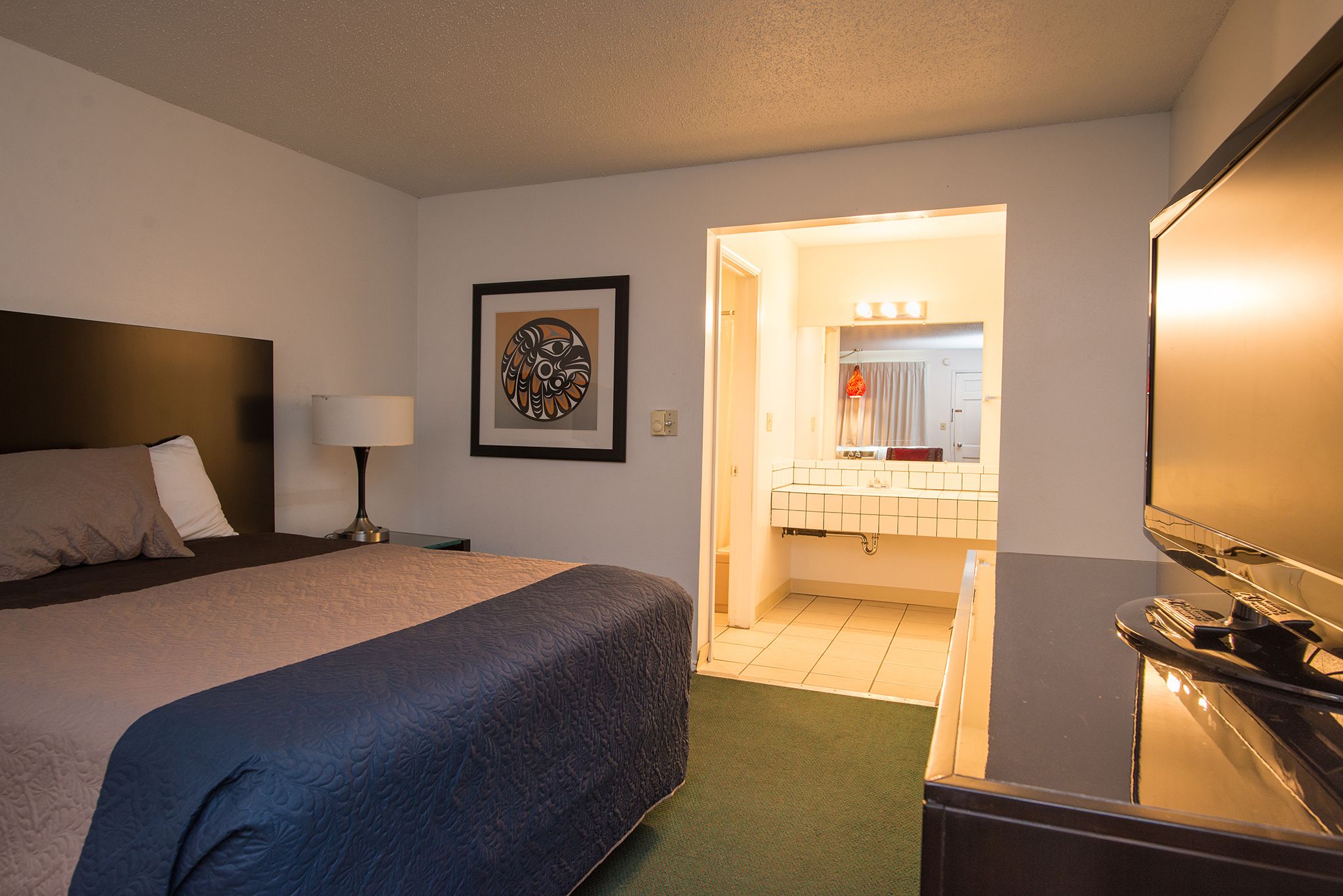 A hotel room with a bed, a desk, a television, a bathroom with a sink and mirror, and a framed artwork on the wall.