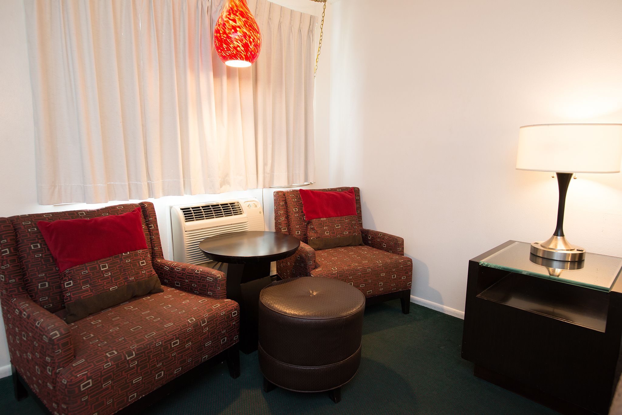 A hotel room with two patterned armchairs, a small round ottoman, a table, a lamp, and a window with curtains.