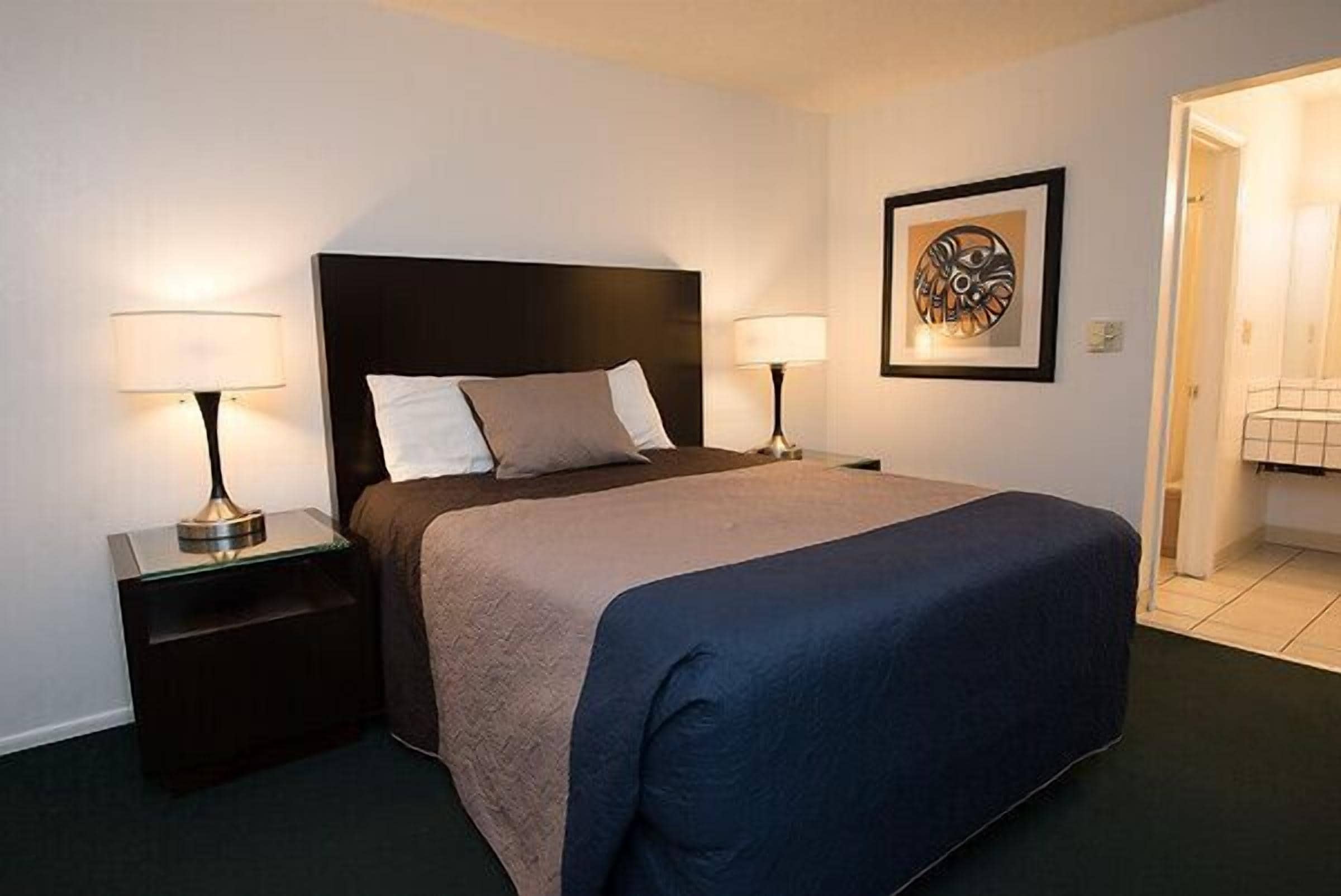 Una habitación de hotel con una cama, dos lámparas de noche, una obra de arte enmarcada en la pared y una puerta que da a otra habitación.