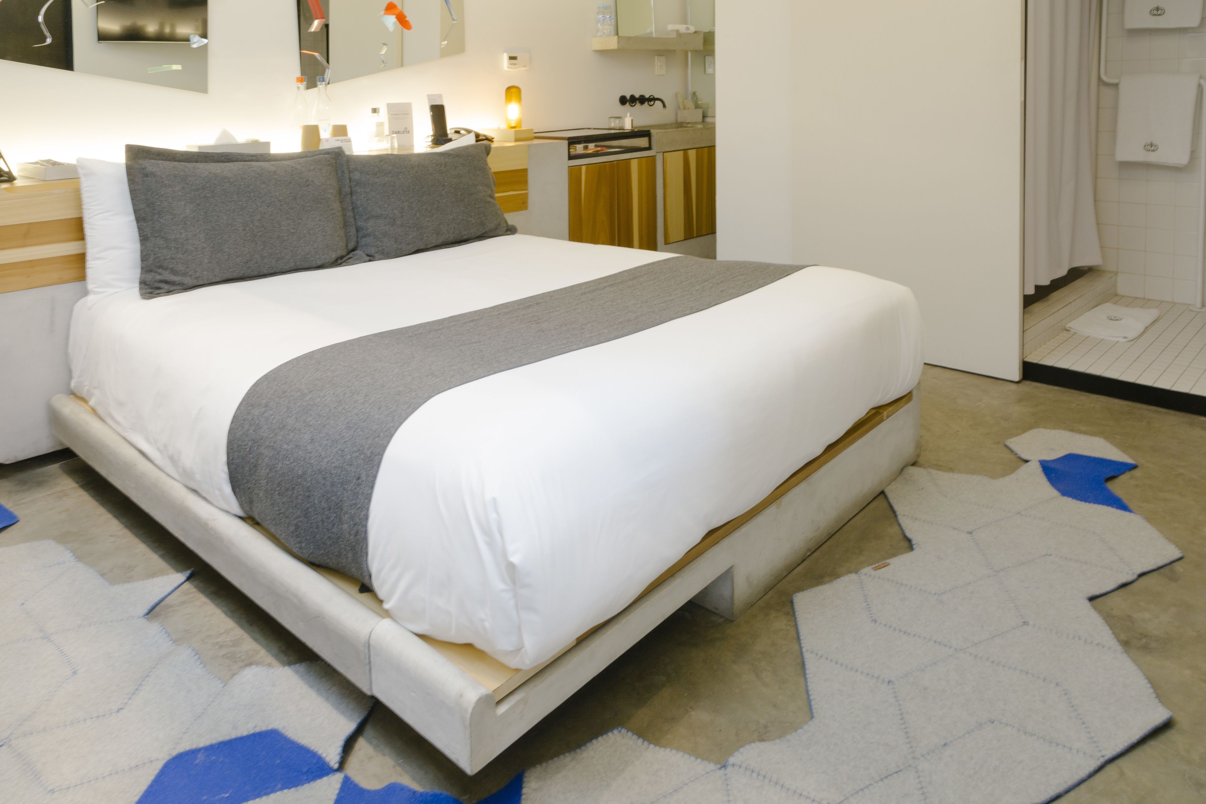 Un lit bien fait, recouvert d’une couverture blanche et grise, avec une tête de lit en bois et une petite table de chevet sur laquelle se trouve une lampe.