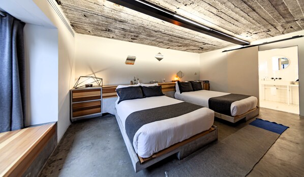 A modern hotel room with two beds, a wooden nightstand, a desk, and a bathroom visible through an open door.
