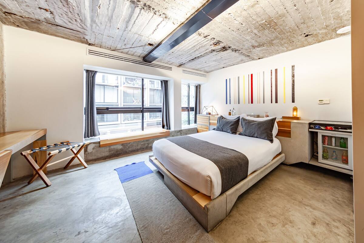 A modern hotel room with a large bed, a desk, a minibar, and a view of the city through the window.