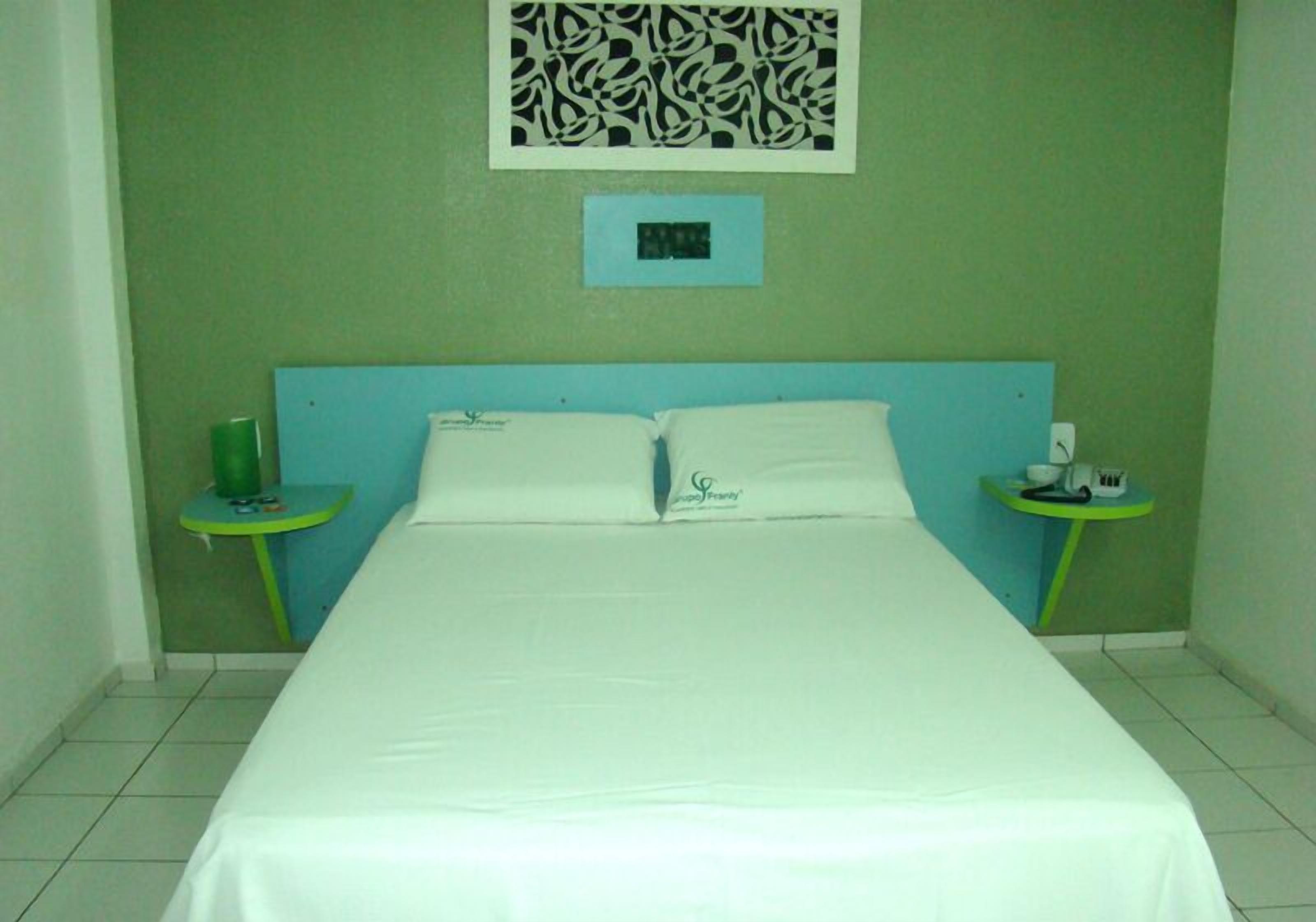 Quarto de hotel com cama, duas mesas de cabeceira, um relógio de parede e um objeto decorativo na parede.