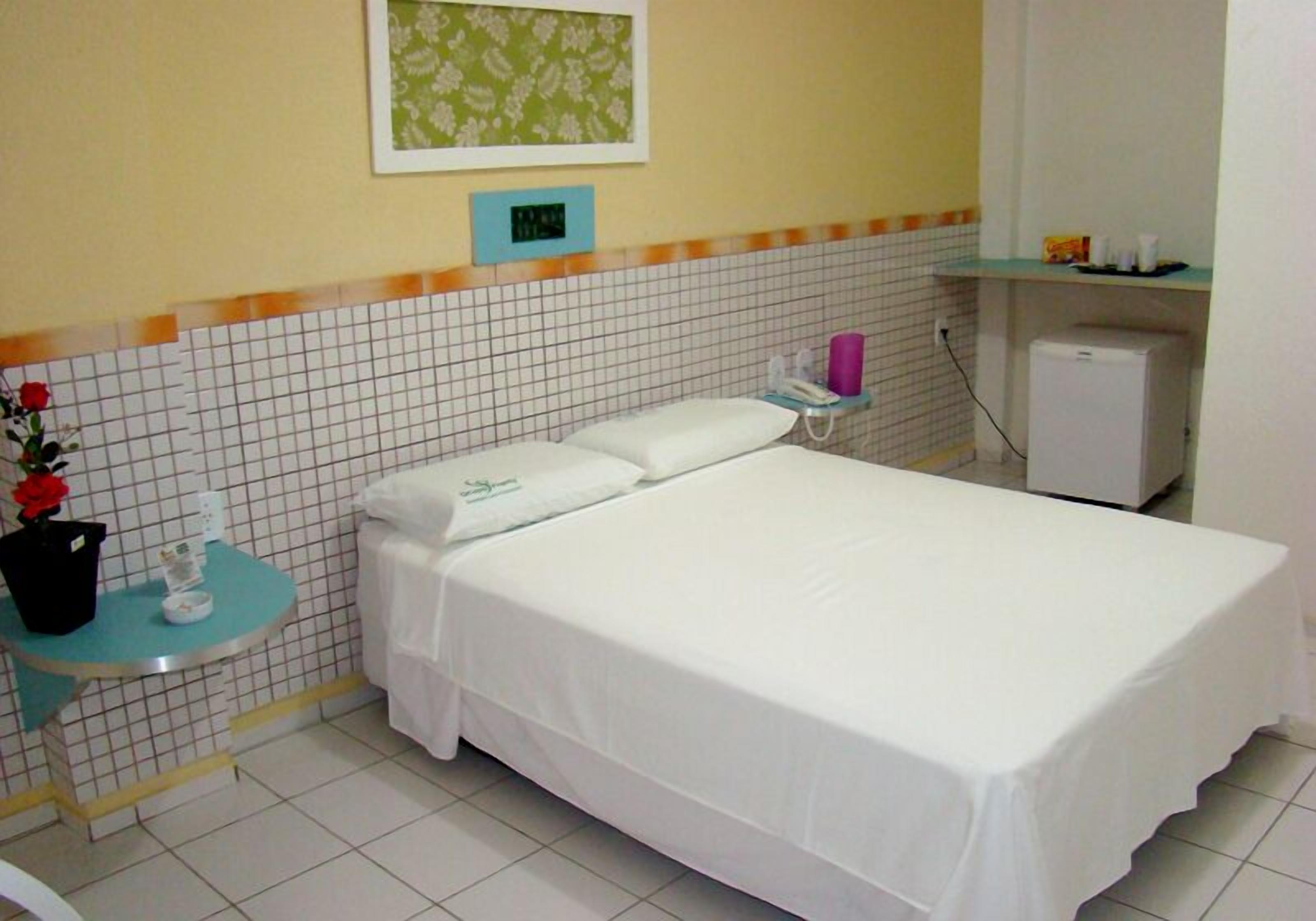 A hotel room with a bed, a small table with a vase of flowers, a telephone, and a refrigerator.