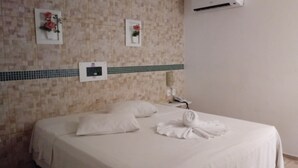 Un cuarto de hotel con una cama bien hecha, un aire acondicionado de pared y dos cuadros con flores en la pared.