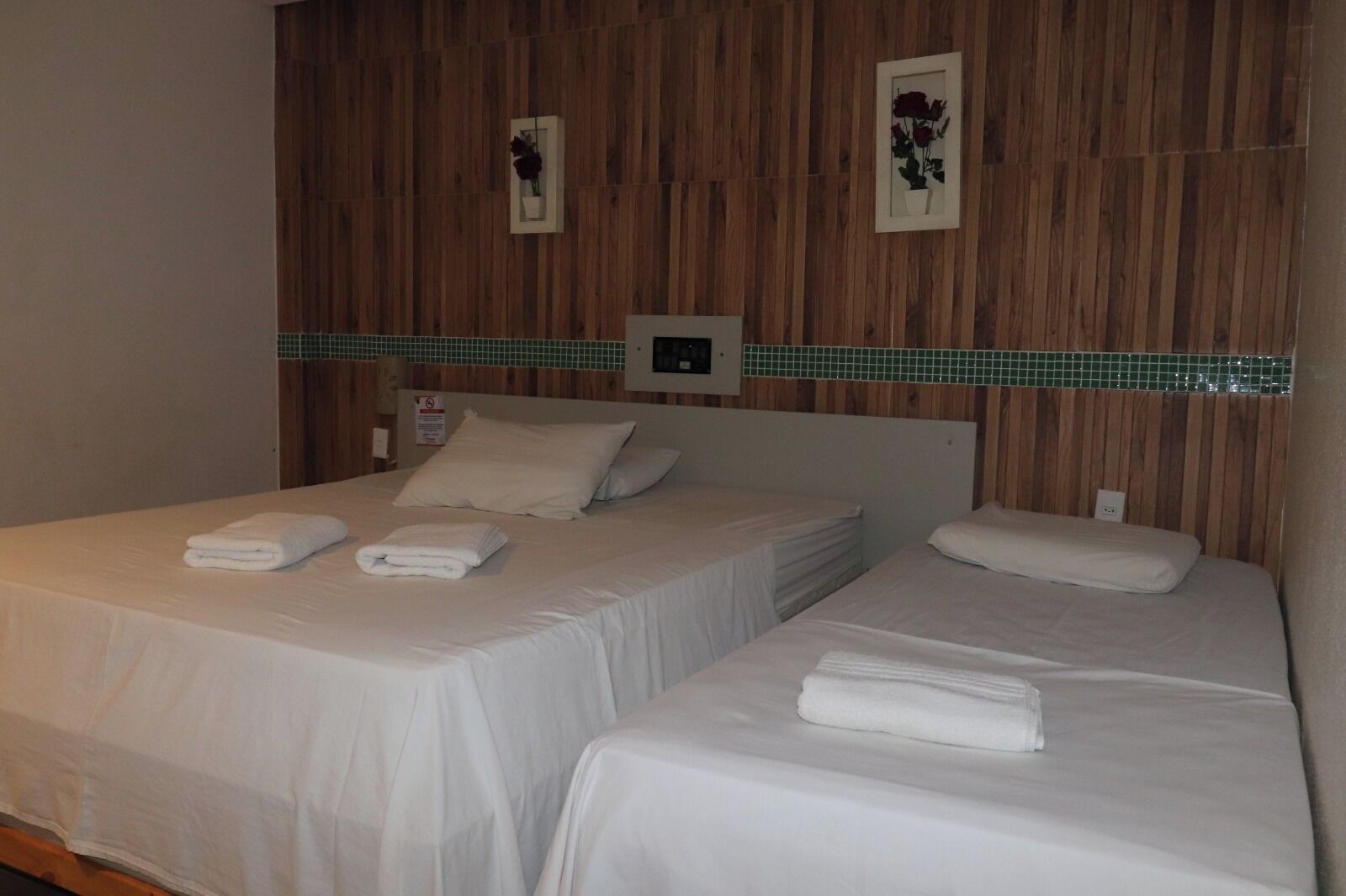 Quarto de hotel com duas camas, cada uma com uma toalha branca dobrada, painéis de madeira nas paredes e uma faixa decorativa em azulejos verdes.